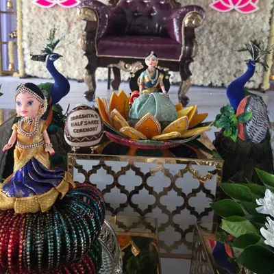A close-up of the decorative elements for a half-saree ceremony, including traditionally dressed dolls, peacock figures, and a personalized coconut.