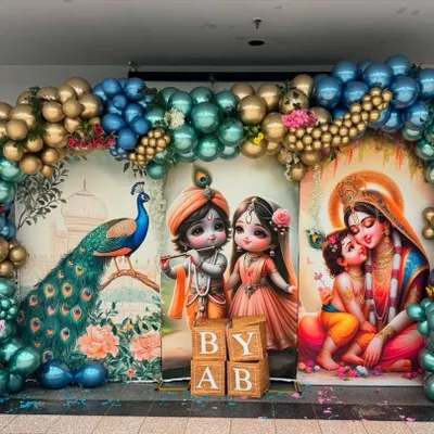 This vibrant baby shower backdrop features three distinct panels with illustrations of a peacock, baby Radha-Krishna, and Yashoda with Krishna. The scene is framed by a large balloon arch in metallic blue and gold, creating a grand and colorful setting.