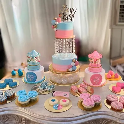 A full dessert table for a "Boy or Girl" themed baby shower. It includes a tiered cake with a crystal separator, smaller carousel cakes, cupcakes, donuts, cakesicles, and heart-shaped treats.