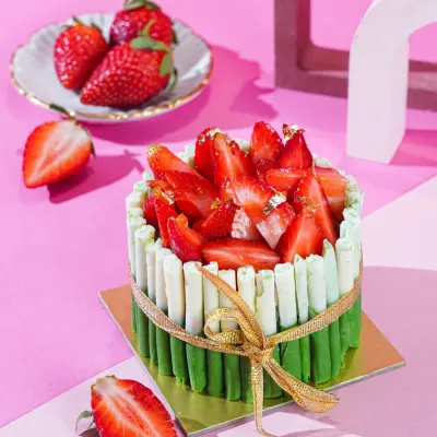 A beautiful strawberry cake with a green and white chocolate fence, tied with a golden ribbon. The bright pink background and modern props create a chic and celebratory feel.