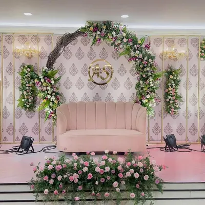 This is the complete stage setup for a rustic-themed engagement. The design is built around a natural twig arch adorned with flowers, a custom gold monogram, and a soft pink sofa, all brought together with warm stage lighting.