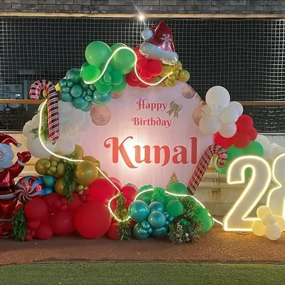 A festive Christmas-themed birthday party for Kunal's 28th, featuring a large personalized backdrop surrounded by red, green, and white balloons. The decor is complete with a Santa Claus balloon, a balloon Christmas tree, candy cane details, and a large light-up number '28'.