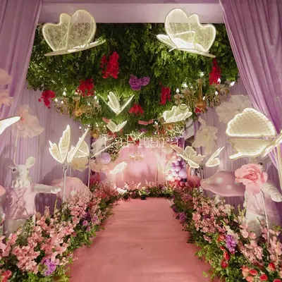 The magical entrance walkway to an enchanted garden party. Guests walk down a pink carpet lined with flowers, whimsical props, and illuminated butterflies hanging from a ceiling of greenery.