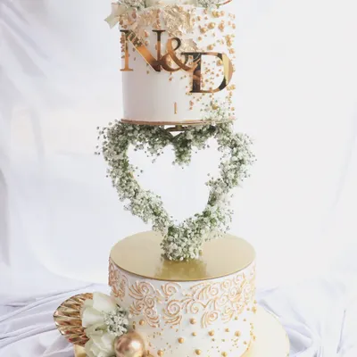 This is one of my favorite engagement cakes, featuring a floating heart-shaped floral wreath. The two tiers are decorated with intricate gold piping and custom initials, making it a stunning centerpiece for a 'Baat Pakki' or engagement party.