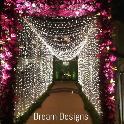 A gorgeous entrance tunnel for a grand wedding. The pathway is framed by an arch of purple roses and walls of cascading fairy lights, leading guests through a magical passage of light and flowers.