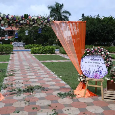 An outdoor event entrance arch decorated with florals and orange fabric. A welcome sign on an easel is also provided as part of the entrance decor rental.