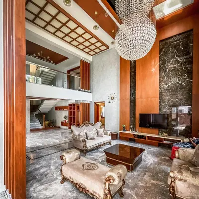 The grand living room of a private villa in Hennur, designed to impress with its double-height volume. A massive crystal chandelier, ornate furniture, and polished marble floors define this opulent space.