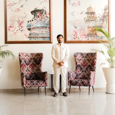 A full-length portrait of the groom, Piyush, in his elegant white engagement sherwani. The symmetrical composition with the large art pieces creates a formal and sophisticated look.