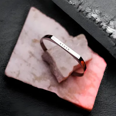 The black-plated cuff bracelet, engraved with 'AYUSH', resting on a piece of pink and white marble. This shot highlights the unique and modern appeal of the black finish.