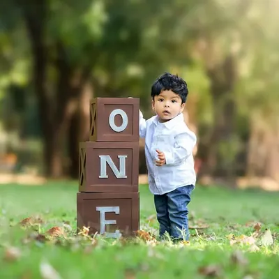 A toddler celebrates his first birthday outdoors, standing next to "ONE" blocks on the green grass of a park.