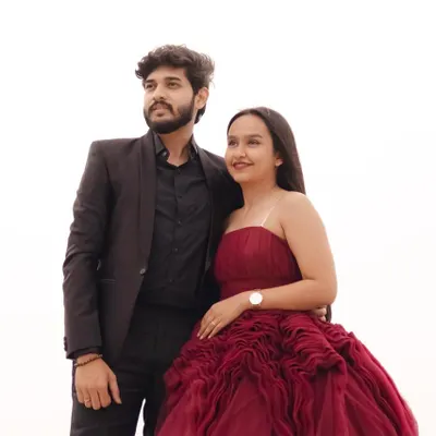 A classic and confident couple's portrait. The bride-to-be wears a stunning strapless maroon gown with intricate bodice detailing, perfectly complemented by her partner's sharp all-black suit.