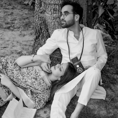 A beautiful black and white portrait capturing a quiet, romantic moment under a tree. The groom holds a vintage camera, and the couple's classic attire adds to the timeless, storybook feel of the image.