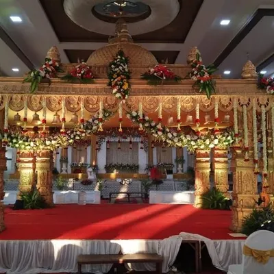 A beautiful and traditional marriage mantapa, featuring ornate golden pillars and a roof adorned with fresh flowers. The red carpet and stage create a classic and regal setting for the wedding rituals.