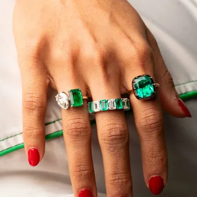 The vibrant green of these emerald rings contrasts beautifully with the crisp white and red nails. This is a powerful lifestyle shot that showcases the luxury and bold style of the jewellery.