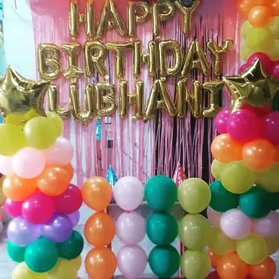 A bright and colorful birthday setup perfect for a home celebration. This decoration for Lubhani features a pink foil curtain, gold "Happy Birthday" letter balloons, and balloon pillars in a rainbow of colors.