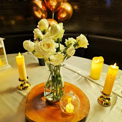 A close-up of an elegant table centerpiece. A simple glass vase with white roses sits on a rustic wood slice, surrounded by the warm glow of safe, battery-operated LED candles in golden holders.