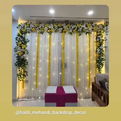 A simple and elegant indoor Haldi backdrop with white drapes, marigold strings, and fairy lights. The setup is completed with a floral border and seating, perfect for home ceremonies.