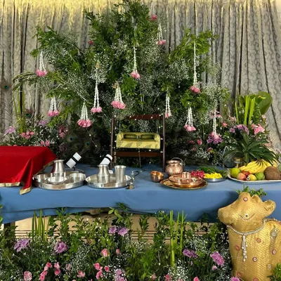 A beautiful Janmashtami altar ready for celebrations. The setup features a miniature swing for Lord Krishna, surrounded by lush greenery, pink flowers, and offerings of fruit, all set against an elegant curtained backdrop.