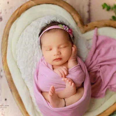 A beautiful 20-day-old baby girl, peacefully asleep. We posed her in a classic womb-like curl within our heart-shaped prop, using a soft purple wrap to add a touch of color.