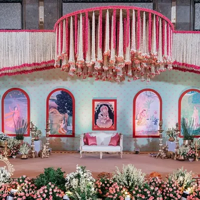 A grand and traditional stage for a South Indian half-saree function. The decor features massive floral chandeliers made of jasmine and pink flowers, with a custom backdrop depicting the stages of a woman's life.