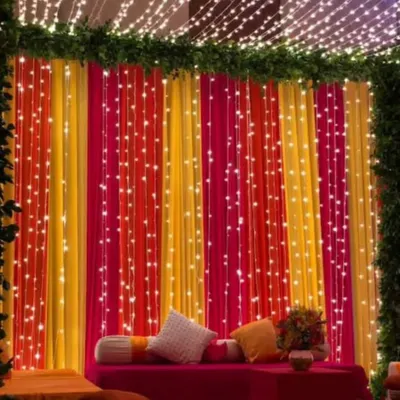 A vibrant backdrop for a Mehendi or Sangeet night, featuring colorful pink, yellow, and orange drapes. The entire setup is illuminated with strings of fairy lights for a magical effect.