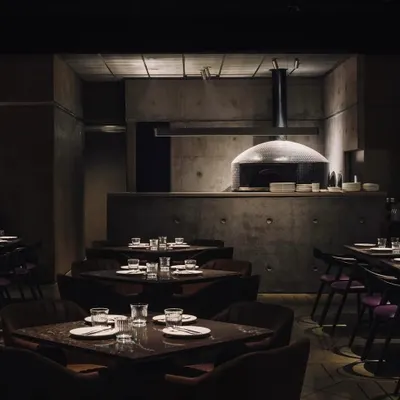 The main dining area at Evoo, where the wood-fired pizza oven serves as a focal point. The space is defined by the monolithic presence of exposed concrete walls and ceilings, creating a dark, atmospheric setting for this pizzeria in Gurugram.