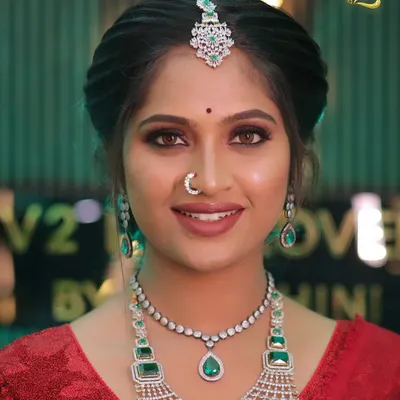 A full view of a stunning modern reception look. The bride is styled in a vibrant red saree, paired with a multi-layered diamond and emerald necklace set. The makeup is glamorous yet fresh, creating a sophisticated look perfect for an evening celebration.