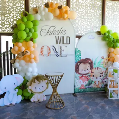 A 'Wild One' jungle theme decoration for a first birthday party. This setup includes a custom backdrop, organic balloon garlands in green, orange, and white, and cute animal cutouts like a lion and elephant to complete the safari look.