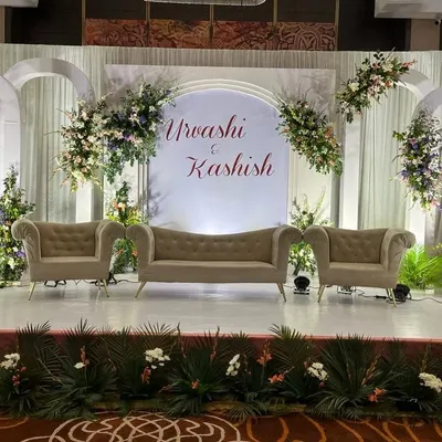 A chic and modern engagement stage design. It features a clean white backdrop with arched panels, the couple's names in elegant calligraphy, and asymmetrical floral arrangements for a sophisticated look.