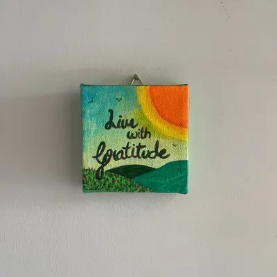"Live with Gratitude." This mini 4x4 inch canvas is a simple, colorful reminder to appreciate the small joys, featuring a bright sun over green hills.