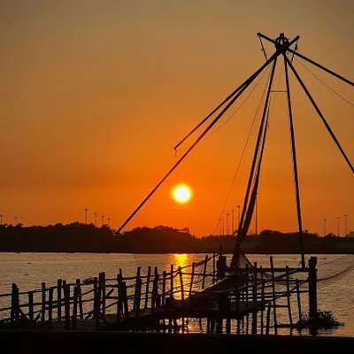 A classic sunset view in Kochi, Kerala, featuring the iconic Chinese fishing net silhouetted against a fiery orange sky. This image captures a unique cultural landscape at the most beautiful time of day.
