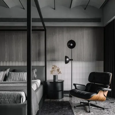 The master bedroom in the Vaulted Haus features a large four-poster bed and an Eames lounge chair. The headboard wall combines backlit frosted glass bricks with grey veneer strips, creating a multi-layered, tactile surface.