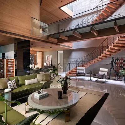This expansive living room in the 'Nadhyavart' project showcases a multi-level design connected by a wooden staircase. The space is unified by a wood-clad ceiling and a skylight that floods the area with natural light, creating a warm and inviting atmosphere.