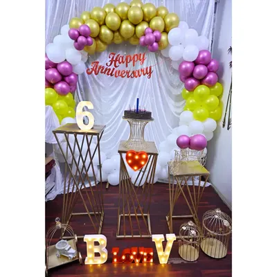 An anniversary party setup with a white, gold, and pink balloon arch. The decoration includes modern gold pedestals, birdcage props, and marquee letters spelling out initials.