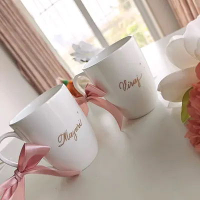 A lovely anniversary gift for a couple. These white ceramic mugs are personalized with the names 'Mayuri' and 'Viraj' in an elegant gold script, tied with pink ribbons for a celebratory finish.