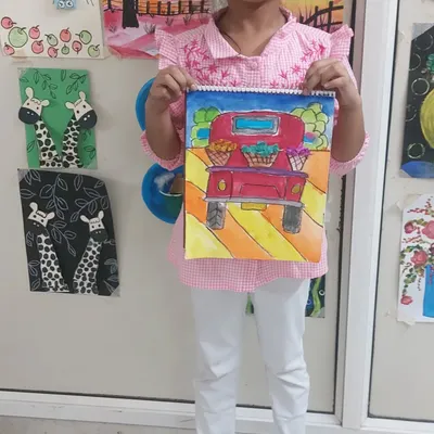 A proud student displays her colorful truck painting. Our art classes guide children through different techniques like watercolor and pattern drawing to create vibrant, detailed artwork.