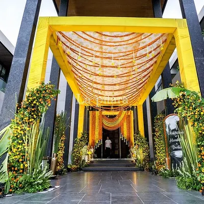 A grand entrance designed for a Teej celebration, featuring a bright yellow canopy adorned with marigold garlands. The pathway is lined with lush greenery and floral arrangements, creating an impressive and welcoming passage for guests.