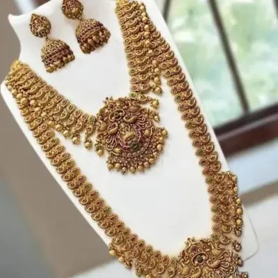 A complete temple jewellery set in antique gold finish, laid out on a display. This set includes a short necklace, a long haram, and matching jhumkas, all featuring intricate peacock and goddess motifs.
