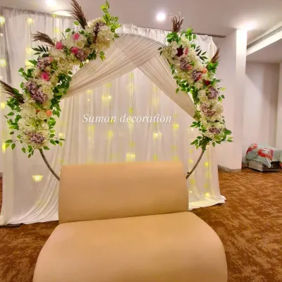 This backdrop features a circular arch decorated with artificial flowers and pampas grass. The white fabric drapes and fairy lights create a soft, glowing background.