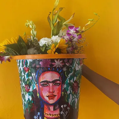 This hand-painted planter features a portrait of Frida Kahlo, surrounded by a lush floral pattern. It's a functional piece of art designed to bring life and color to any room or balcony.