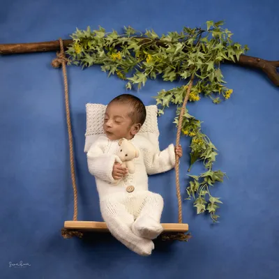 Swinging through life with a heart full of dreams. This baby is sleeping peacefully on a miniature swing, holding a teddy bear, set against a beautiful blue sky backdrop.