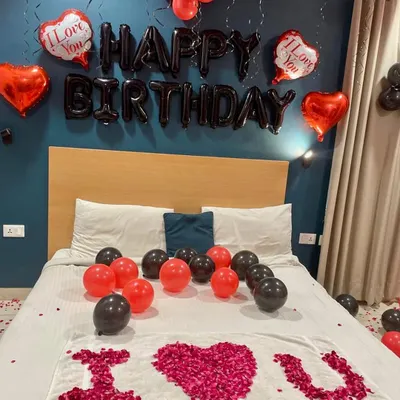 A classic romantic birthday room decoration. This setup includes "Happy Birthday" foil balloons, red and black latex balloons, and a heart-shaped "I Love You" message created with rose petals on the bed.