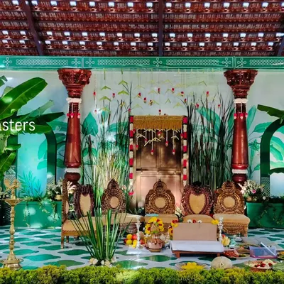 A closer view of the traditional wedding mandap, focusing on the seating area for the couple. The decor features carved wooden chairs, brass lamps, and a backdrop of hand-painted greenery and hanging floral strings, all under a classic tiled roof.