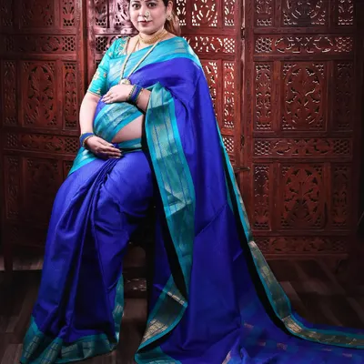 A regal portrait of a mother-to-be in a stunning blue and turquoise silk saree. The traditional jewelry and pose against a carved wooden screen create a timeless and culturally rich image.