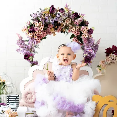 A beautiful portrait for a first birthday celebration. This little girl is all smiles in her fluffy purple and white dress, sitting on a miniature couch under a floral arch.