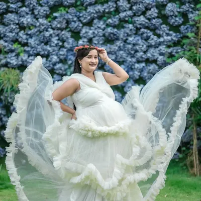 A full-length view of our popular white ruffled gown against a beautiful wall of blue flowers. This outdoor setup at our studio is perfect for creating dramatic and angelic maternity portraits.