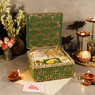 An open view of our green and gold woven Kauna box, a highlight of our Diwali collection. It's filled with festive goodies, including a traditional brass diya, sweets, and fragrant items, making it a complete sensory experience.