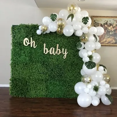A clean and classic 'Oh Baby' backdrop. This setup uses a lush green hedge wall as the base, with a cascade of white and gold confetti balloons and tropical leaves for a fresh, botanical feel.