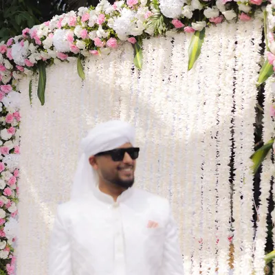 The groom looking cool and confident in his all-white Nikaah outfit, complete with sunglasses. A stylish portrait against a beautiful floral backdrop.