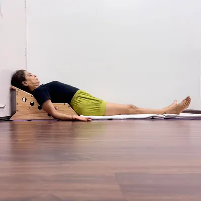 This is a supported Savasana on a Simhasana Box, a prop designed to open the chest and support the spine's natural curves. This posture encourages deeper breathing and helps the body shift into a state of rest and recovery.